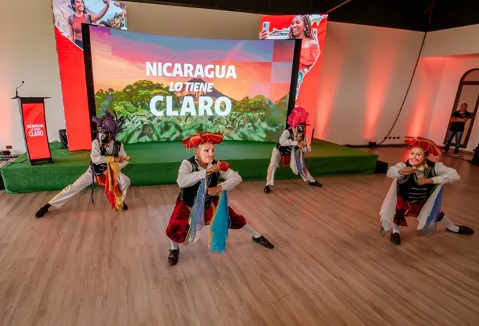 Cuatro bailarines con trajes y máscaras tradicionales actúan en un escenario frente a una pantalla grande con texto.