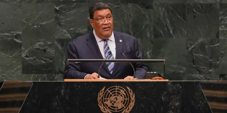 Un hombre con traje y corbata, con gafas, habla en un podio frente a un fondo de mármol verde. El emblema de las Naciones Unidas es visible en el podio.