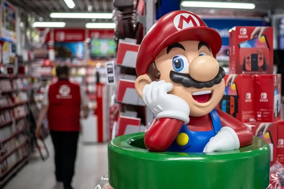 Figura grande del personaje Mario, con su icónico gorro rojo y overol azul, en primer plano dentro de una tienda de electrónica. Al fondo, estanterías con cajas de videojuegos y consolas, y un empleado con chaleco rojo.