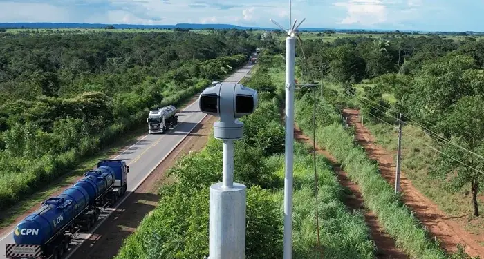 Cámara de vigilancia en poste metálico supervisando el tráfico de camiones cisterna en una carretera rodeada de vegetación.