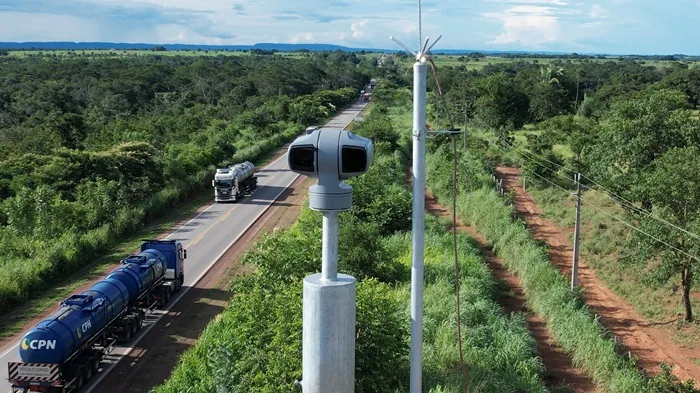 Cámara de vigilancia en poste metálico supervisando el tráfico de camiones cisterna en una carretera rodeada de vegetación.