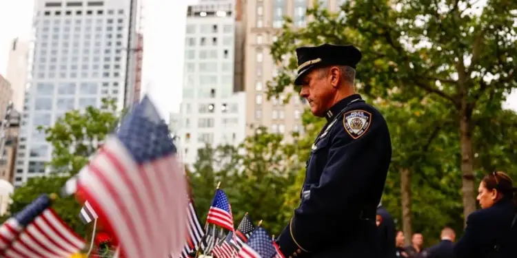 Oficial de policía de Nueva York con uniforme azul oscuro, gorra y parche en el hombro, mirando hacia abajo. Banderas de EE. UU. y flores amarillas en primer plano. Edificios altos y árboles al fondo.