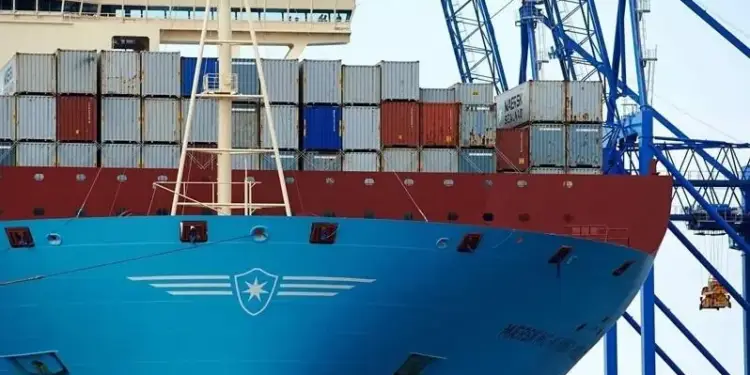 Primer plano de la proa de un gran buque portacontenedores azul y rojo, cargado con múltiples contenedores apilados, junto a grúas portuarias azules.