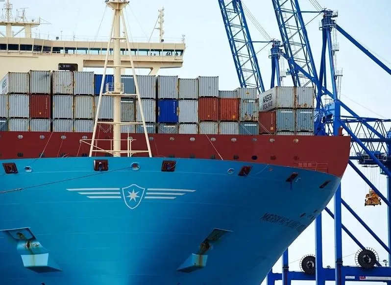 Primer plano de la proa de un gran buque portacontenedores azul y rojo, cargado con múltiples contenedores apilados, junto a grúas portuarias azules.