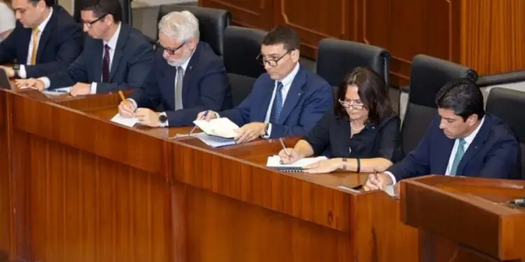 Grupo de seis profesionales, cinco hombres y una mujer, sentados en una mesa de madera oscura, tomando notas y revisando documentos durante una reunión de negocios formal. Visten trajes y camisas, mostrando un ambiente de trabajo concentrado.