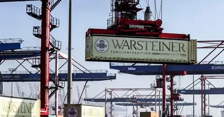Grúas portuarias operando en un muelle, levantando y moviendo contenedores de carga con la marca Warsteiner y el texto 'Familientradition seit 1753'. Varios contenedores están en trenes en las vías.