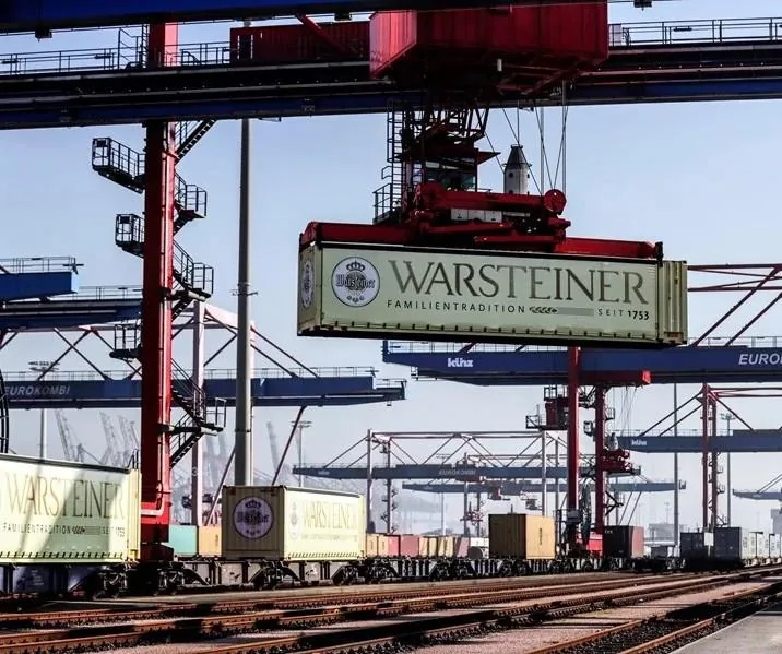 Grúas portuarias operando en un muelle, levantando y moviendo contenedores de carga con la marca Warsteiner y el texto 'Familientradition seit 1753'. Varios contenedores están en trenes en las vías.