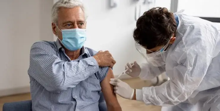 Un hombre mayor con mascarilla recibe una inyección en el brazo de un profesional de la salud con equipo de protección, simbolizando la inmunización.