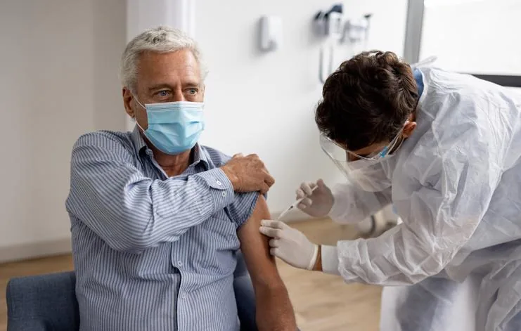 Un hombre mayor con mascarilla recibe una inyección en el brazo de un profesional de la salud con equipo de protección, simbolizando la inmunización.
