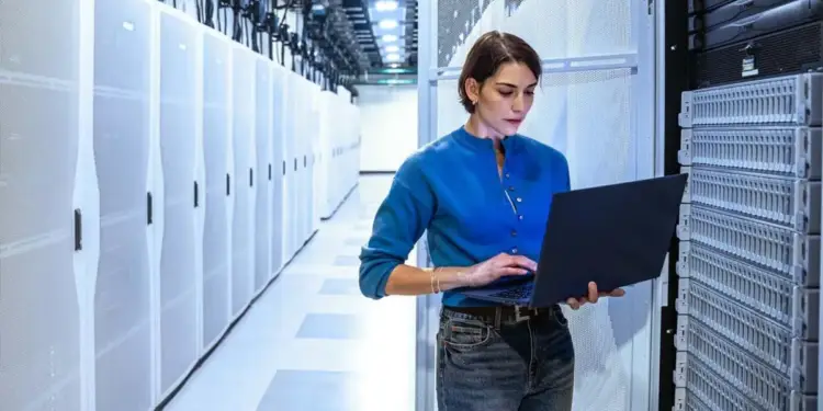 Mujer profesional con camisa azul y jeans, usando una laptop en un moderno centro de datos. Se aprecian filas de servidores y racks.