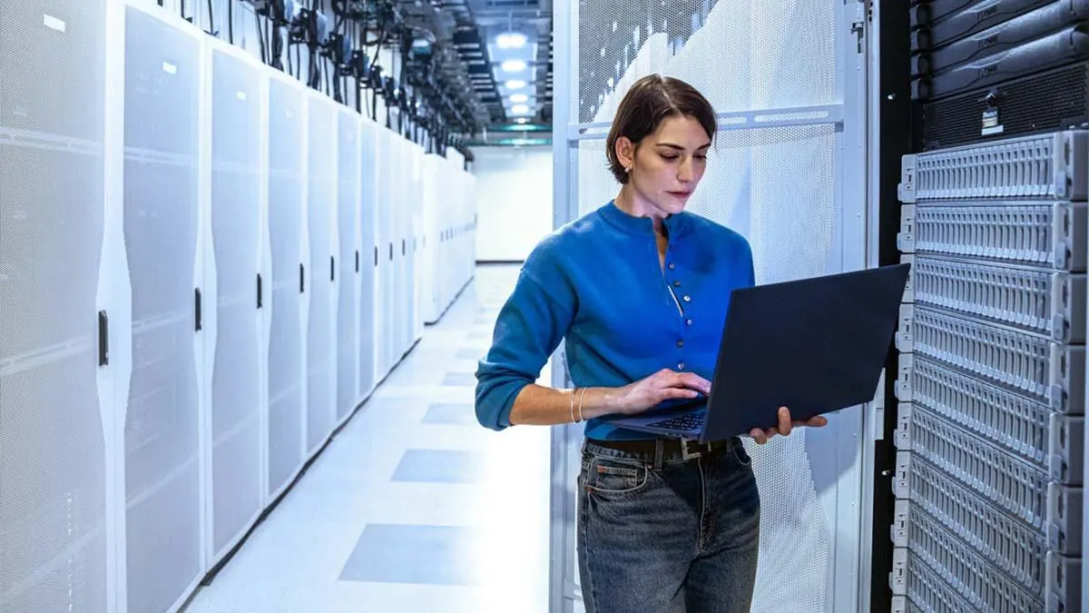 Mujer profesional con camisa azul y jeans, usando una laptop en un moderno centro de datos. Se aprecian filas de servidores y racks.