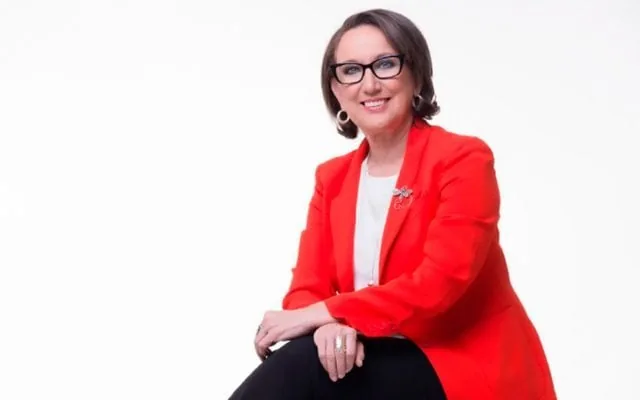 Mujer profesional sonriendo, vistiendo chaqueta roja y gafas, sentada sobre fondo blanco.