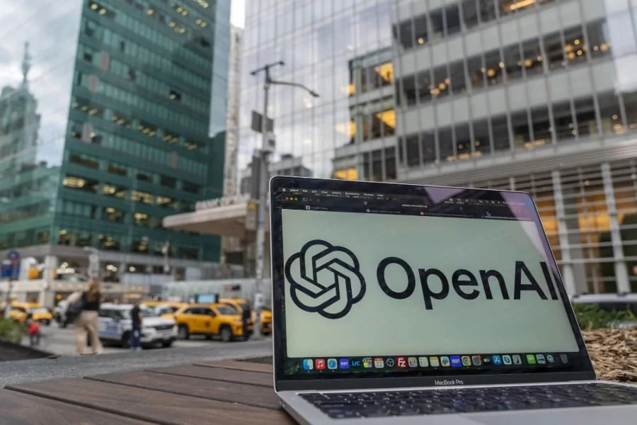 Primer plano de una laptop mostrando el logo de OpenAI, con edificios de oficinas modernos y taxis amarillos en el fondo, representando el entorno empresarial y tecnológico.