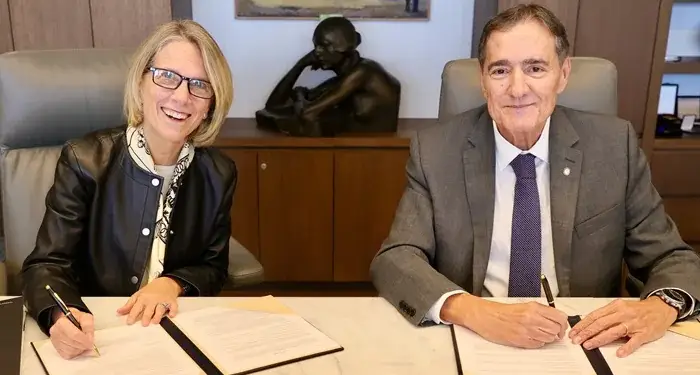 Una mujer con gafas y chaqueta negra y un hombre con traje y corbata firmando documentos en un escritorio de oficina.