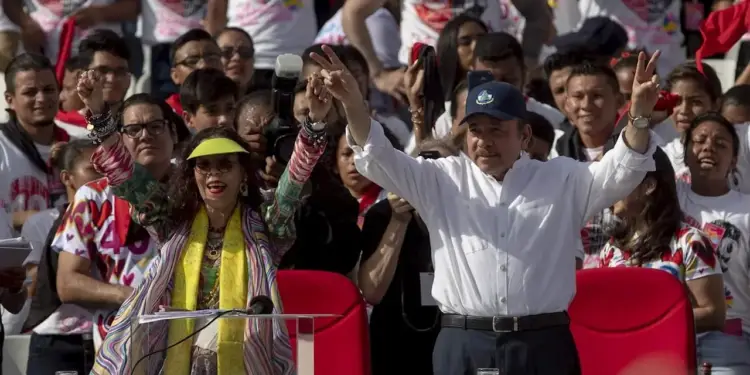 Hombre y mujer con la mano levantada haciendo la señal de la victoria frente a una multitud en un evento.