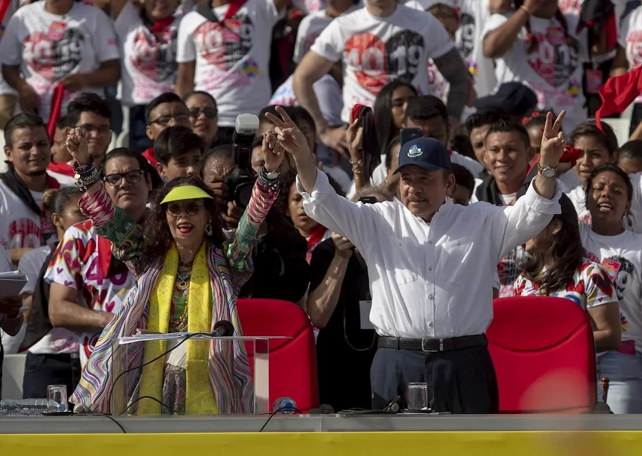 Hombre y mujer con la mano levantada haciendo la señal de la victoria frente a una multitud en un evento.