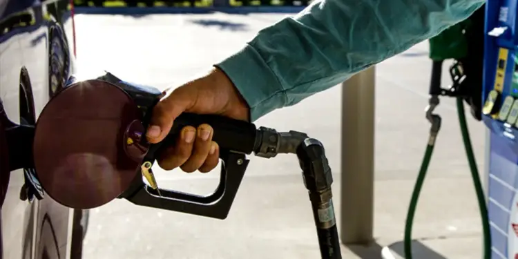 Mano de persona insertando una boquilla de combustible en el tanque de un coche oscuro en una gasolinera. Se observa la manguera verde y parte del surtidor.