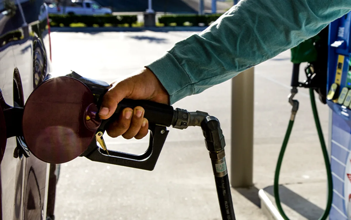 Mano de persona insertando una boquilla de combustible en el tanque de un coche oscuro en una gasolinera. Se observa la manguera verde y parte del surtidor.