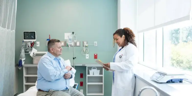 Doctora con bata blanca sonriendo mientras revisa documentos junto a un paciente en un consultorio médico.