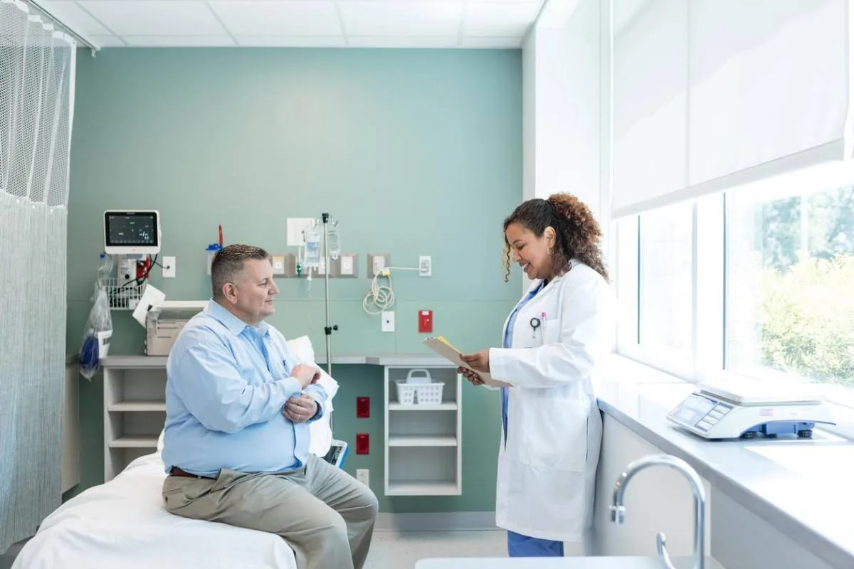 Doctora con bata blanca sonriendo mientras revisa documentos junto a un paciente en un consultorio médico.