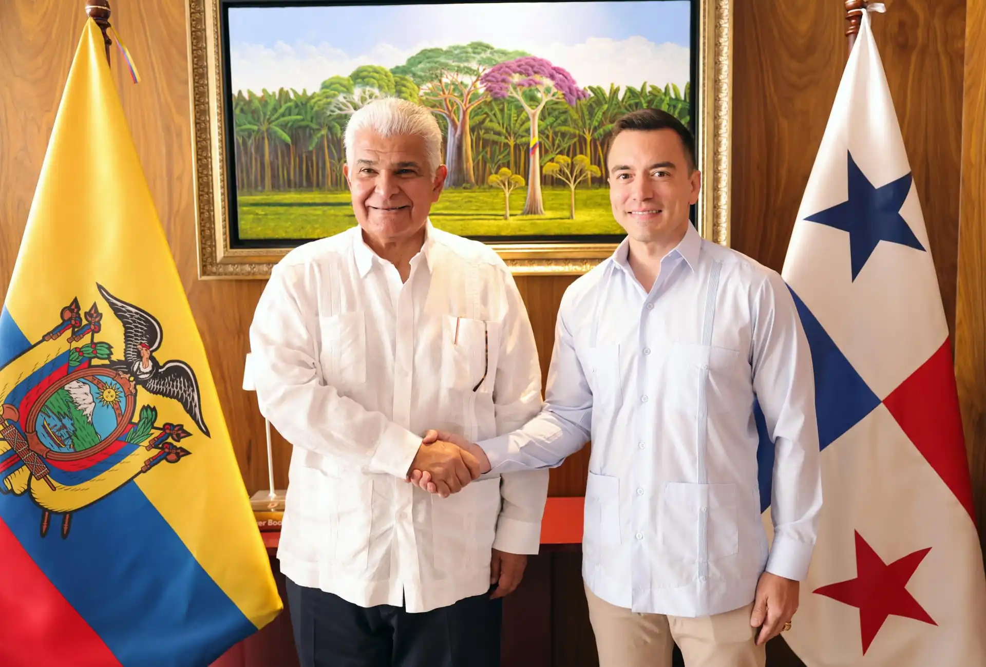 Dos hombres con camisas formales estrechan sus manos frente a banderas institucionales y una pintura al óleo.