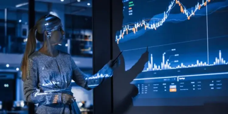 Mujer profesional rubia con coleta señalando un gráfico financiero en una pantalla grande y oscura, mostrando datos bursátiles y análisis.