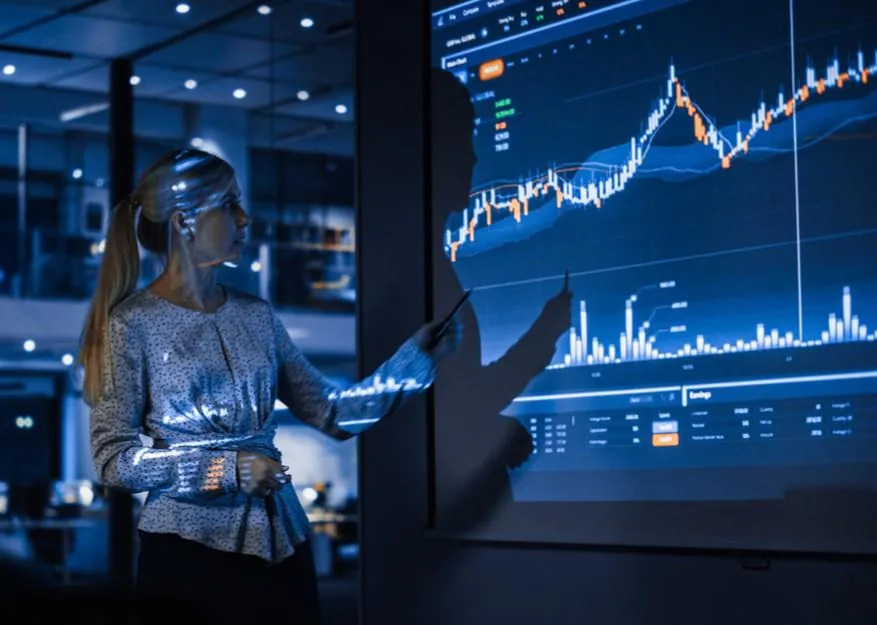 Mujer profesional rubia con coleta señalando un gráfico financiero en una pantalla grande y oscura, mostrando datos bursátiles y análisis.