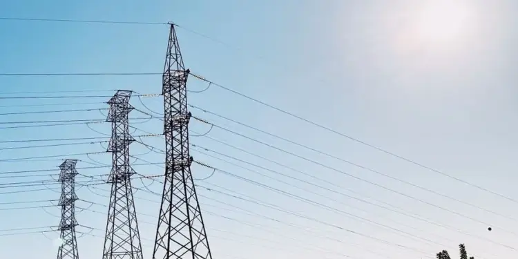 Varias torres de alta tensión de metal se extienden bajo un cielo azul claro con cables eléctricos. Montañas y vegetación en el fondo.