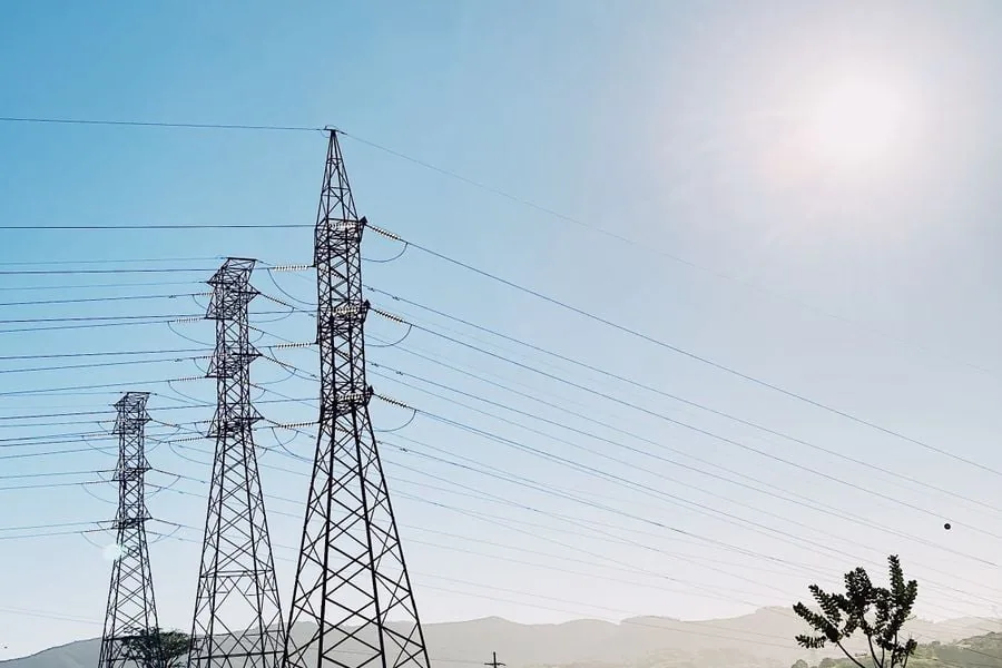 Varias torres de alta tensión de metal se extienden bajo un cielo azul claro con cables eléctricos. Montañas y vegetación en el fondo.