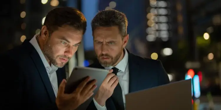Dos hombres de negocios con traje revisando información en una tablet y una laptop en una calle iluminada por la noche.
