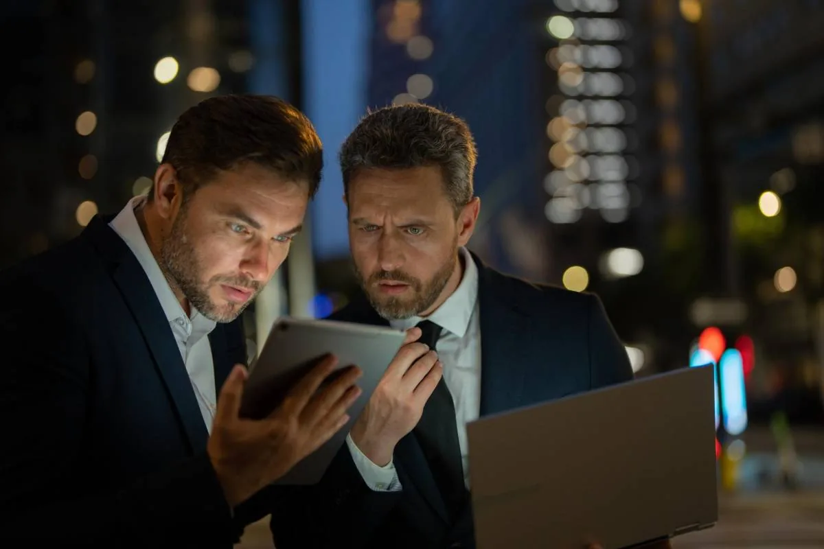 Dos hombres de negocios con traje revisando información en una tablet y una laptop en una calle iluminada por la noche.