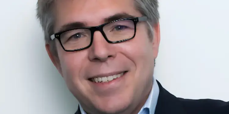 Hombre sonriente con gafas y traje en un retrato profesional sobre fondo claro.