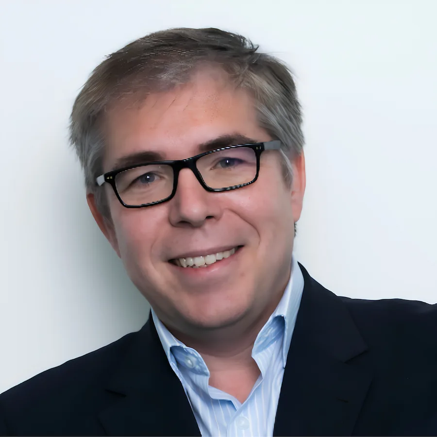 Hombre sonriente con gafas y traje en un retrato profesional sobre fondo claro.