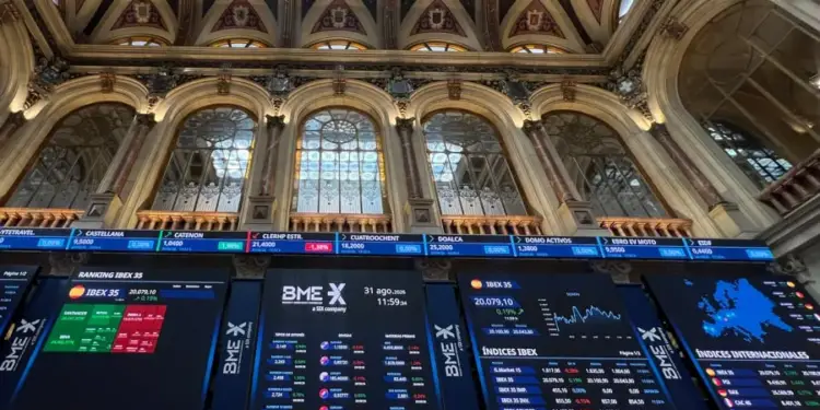 Pantallas digitales muestran datos del IBEX 35 y otros índices bursátiles en un edificio histórico con techos altos y ventanas arqueadas.