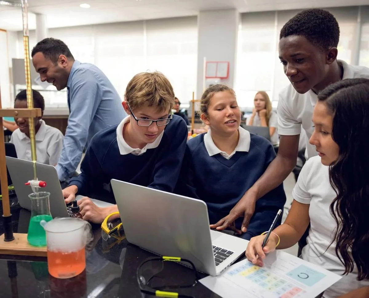 Estudiantes y adultos colaboran en un laboratorio, usando laptops y equipos científicos. Foco en el aprendizaje interactivo y tecnológico.