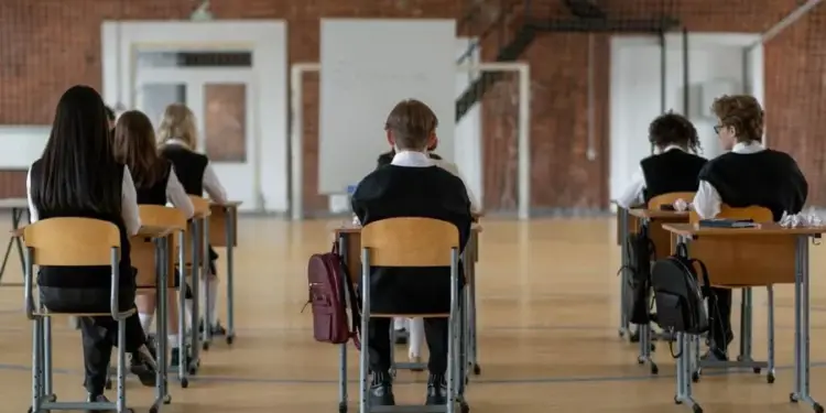 Vista trasera de varios estudiantes sentados en pupitres individuales dentro de un gran salón escolar con piso de madera.