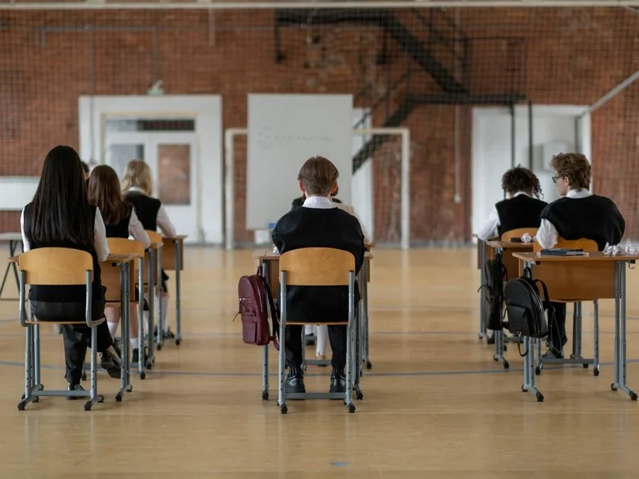 Vista trasera de varios estudiantes sentados en pupitres individuales dentro de un gran salón escolar con piso de madera.
