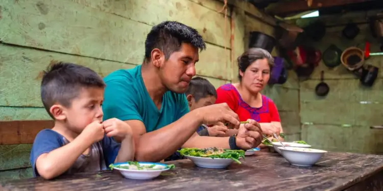 Una familia con dos niños y dos adultos compartiendo una comida sencilla alrededor de una mesa de madera en una vivienda rural.