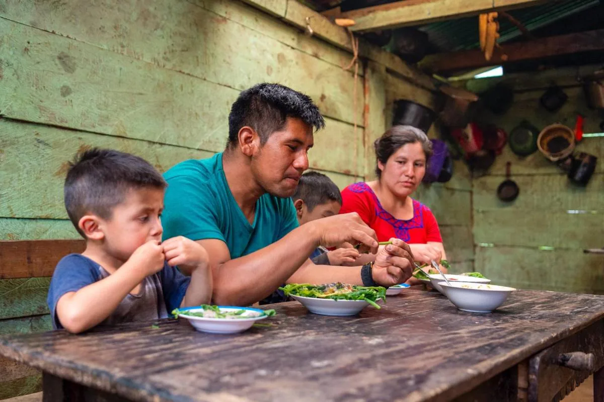Una familia con dos niños y dos adultos compartiendo una comida sencilla alrededor de una mesa de madera en una vivienda rural.