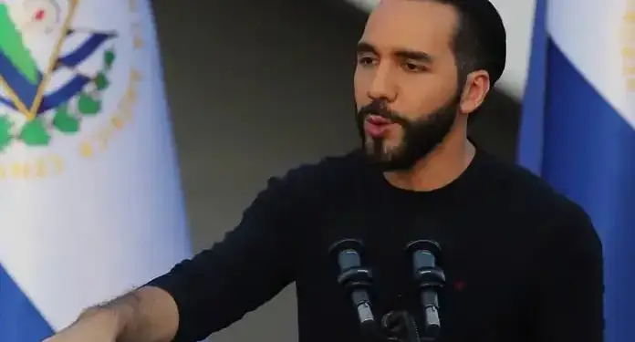 Hombre con barba y ropa negra gesticula al hablar frente a micrófonos con la bandera de El Salvador de fondo.