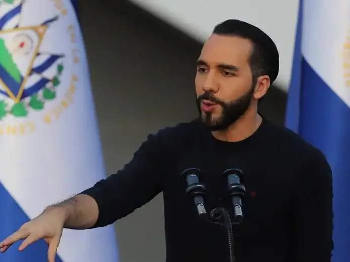 Hombre con barba y ropa negra gesticula al hablar frente a micrófonos con la bandera de El Salvador de fondo.