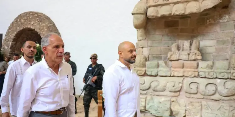 Dos hombres vestidos con camisas blancas y pantalones caminan cerca de una estructura de piedra antigua, acompañados por personal de seguridad con equipo militar.