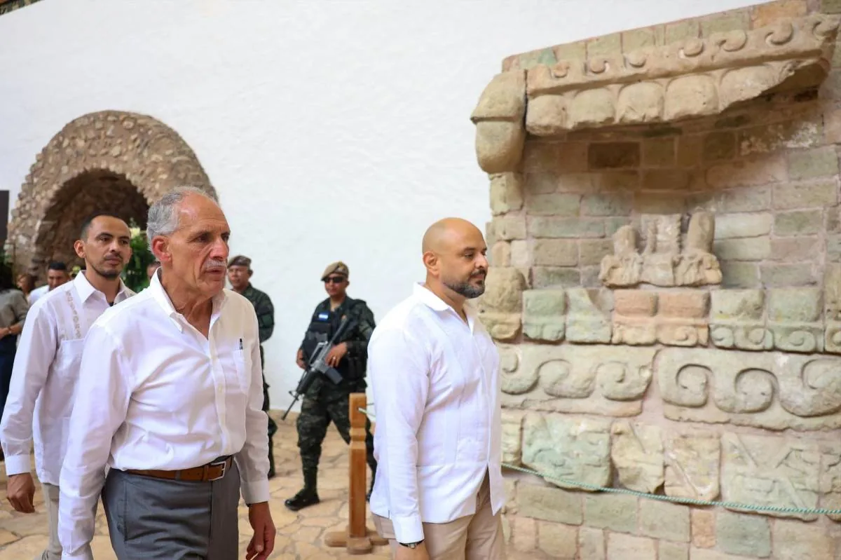 Dos hombres vestidos con camisas blancas y pantalones caminan cerca de una estructura de piedra antigua, acompañados por personal de seguridad con equipo militar.