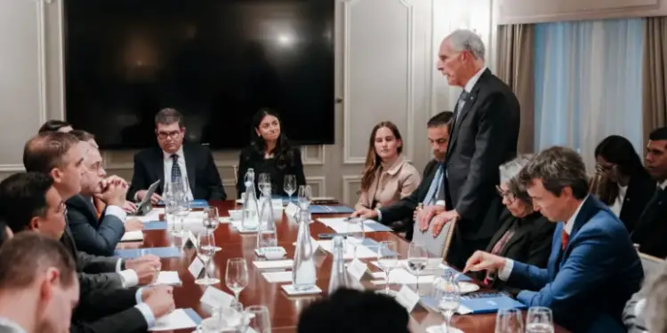 Reunión de negocios con profesionales sentados alrededor de una mesa de madera. Un hombre de pie se dirige al grupo, con una pantalla grande al fondo.