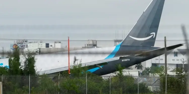 Vista lateral de la cola de un avión de carga Amazon con el logo visible, parcialmente oculto por vegetación y una valla, con edificios industriales al fondo.