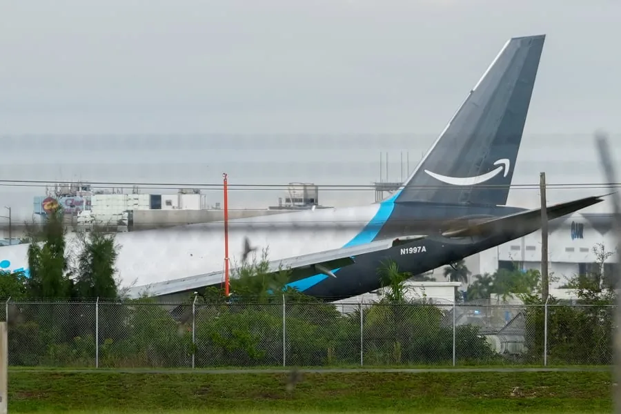 Vista lateral de la cola de un avión de carga Amazon con el logo visible, parcialmente oculto por vegetación y una valla, con edificios industriales al fondo.