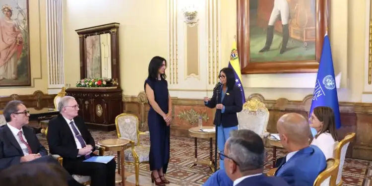 Una mujer de pie habla ante un grupo de personas sentadas en un salón protocolar con banderas y cuadros.