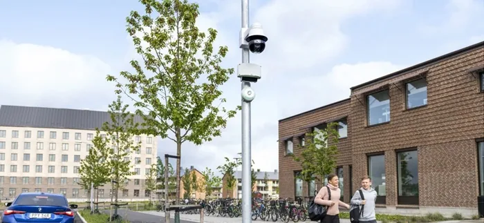 Cámara de vigilancia instalada en un poste metálico en una calle urbana con edificios de ladrillo y peatones caminando.