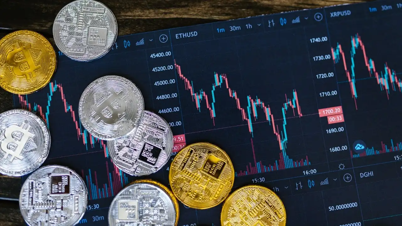 Monedas físicas de criptomonedas doradas y plateadas sobre una pantalla oscura con gráficos de trading bursátil.