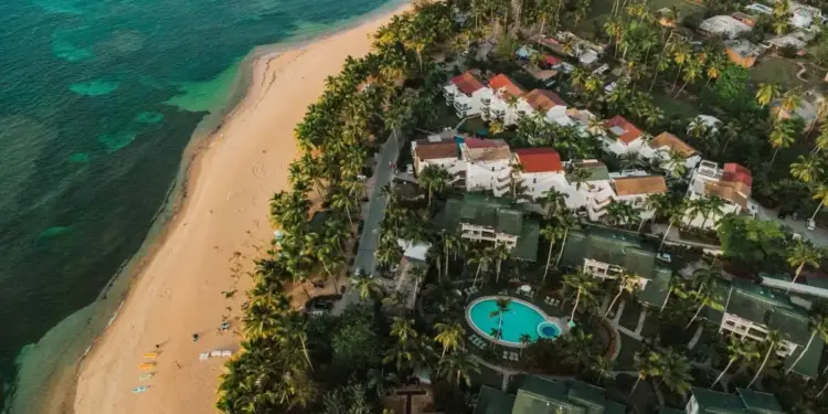 Vista aérea de una playa tropical con arena dorada, mar turquesa, palmeras y complejos turísticos con piscinas.